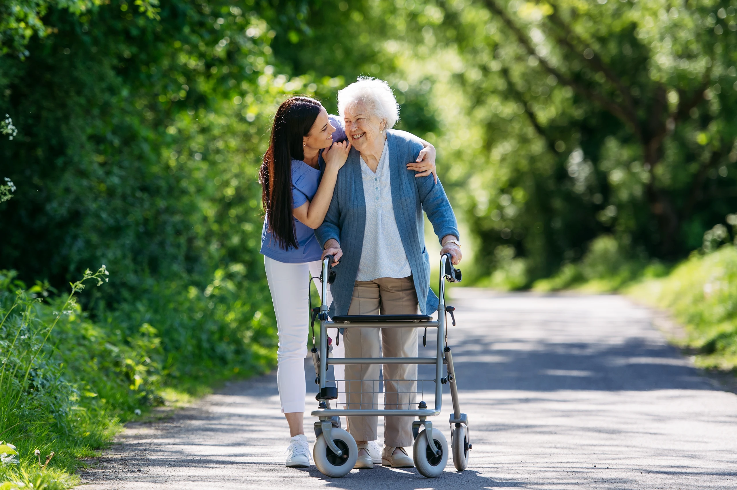 Ältere Frau mit Rollator geht mit jüngerer Frau lachend einen Waldweg entlang.